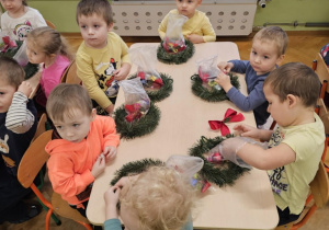 Grupa dzieci siedzi wokół stołu. Na stole leżą wianki i ozdoby w torebeczkach.