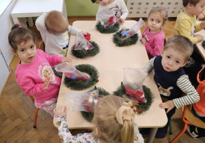 Grupa dzieci siedzi wokół stołu. Na stole leżą wianki i ozdoby w torebeczkach.