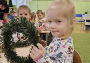 Dziewczynka w jasnych włoskach siedzi przy stoliku i trzyma w dłoni wianek.