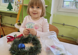 Dziewczynka uśmiecha się do zdjęcia. Na stole leży wianek oraz ozdoby w woreczku.