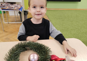 Chłopiec w czarno-szarej bluzce uśmiecha się do zdjęcia. Na stole leży wianek oraz ozdoby w woreczku.