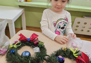 Dziewczynka w jasnej bluzce siedzi przy stoliku. Na stoliku leżą dwa wianki.