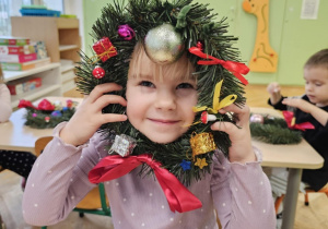 Dziewczynka uśmiecha się do zdjęcia. W dłoniach trzyma wianek.