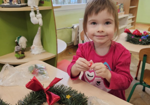 Dziewczynka w różowje bluzce siedzi przy stoliku. Na stoliku leżą dwa wianki.