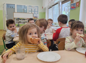 Dzieci siedzą przy stolikach i jedzą pączki. Na pierwszym planie dwie dziewczynki. Jedna ma bluzkę w paski a druga w motylki.