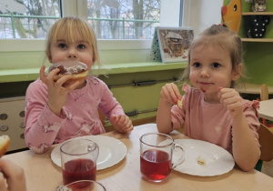 Dwie dziewczynki ubrane na różowo siedzą przy stoliku jedzą pączki.