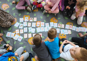Na dywanie w sowy leżą karty. Wokół kart siedzą dzieci.