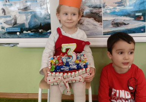 Dziewczynka siedzi na krześle w koronie. W dłoni trzyma tort ze słodyczy.