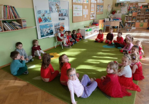 Grupa dzieci siedzi na zielonym dywanie. Jedna z dziewczynek siedzi na krześle w koronie. W dłoni trzyma tort ze słodyczy.