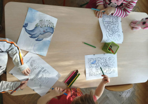 dzieci siedzą przy stoliku i kolorują kolorowanki z okazji Dnia Babci i Dziadka