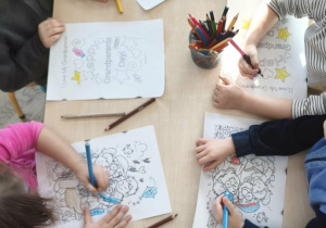 dzieci siedzą przy stoliku i kolorują kolorowanki z okazji Dnia Babci i Dziadka