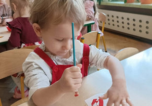 Dziewczynka w białej bluzce i czerwonej sukience siedzi przy stoliku i maluje serce farbami.