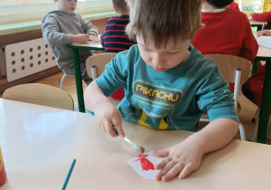 Chłopiec w turkusowej bluzce siedzi przy stoliku i maluje serce farbami.