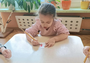 Dziewczynka w różowej sukience siedzi przy stoliku i maluje serce farbami.