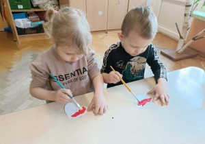 Dziewczynka i chłopiec siedzą przy stoliku i malują serca farbami.