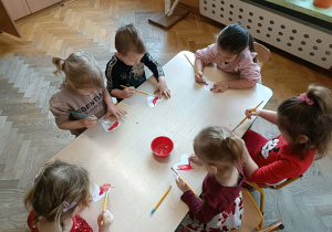 Grupa dzieci siedzi przy stoliku i maluje serca farbami.