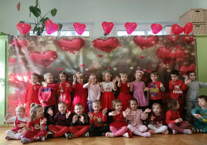 Grupa dzieci stoi na tle szarej dekoracji, na której widać serca.