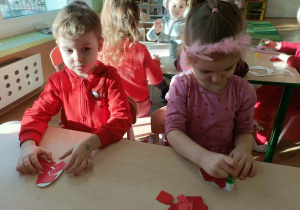 Dwójka dzieci siedzi przy stoliku i wykleja serca skrawkami papieru.