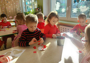 Dwójka dzieci siedzi przy stoliku i wykleja serca skrawkami papieru.