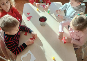 Grupa dzieci siedzi przy stoliku i wykleja serca skrawkami papieru.