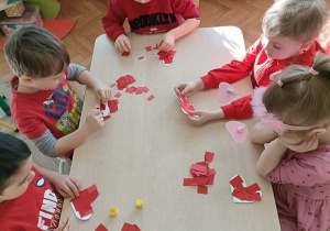 Grupa dzieci siedzi przy stoliku i wykleja serca skrawkami papieru.