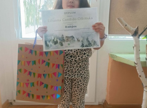 Dziewczynka stoi z dyplomem i nagrodą w ręce. Za nią okno.