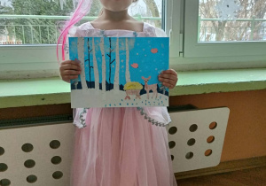 Dziewczynka stoi w różowej sukience i pracą w ręce. Za nią okno.