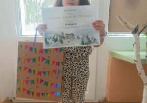 Dziewczynka stoi z dyplomem i nagrodą w ręce. Za nią okno.