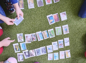 Na zielonym dywanie leżą kolorowe karty. Wokół widać nogi siedzących dzieci.