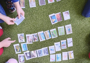 Na zielonym dywanie leżą kolorowe karty. Wokół widać nogi siedzących dzieci.
