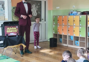 Iluzjonista i dziewczynka w bordowych spodniach stoją na scenie. Po lewo widać kawałek zielonego dywanu.