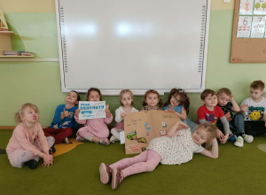 Grupa dzieci siedzi na zielonym dywanie. Trzy dziewczynki trzymają plakat, jedna dziewczynka trzyma kartkę z napisem Dzień Dentysty