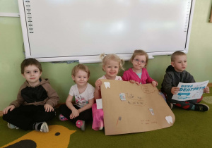 Grupa dzieci siedzi na zielonym dywanie. Dwie dziewczynki trzymają plakat, jeden chłopiec trzyma kartkę z napisem Dzień Dentysty