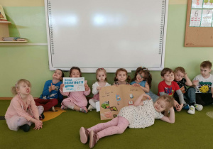 Grupa dzieci siedzi na zielonym dywanie. Trzy dziewczynki trzymają plakat, jedna dziewczynka trzyma kartkę z napisem Dzień Dentysty