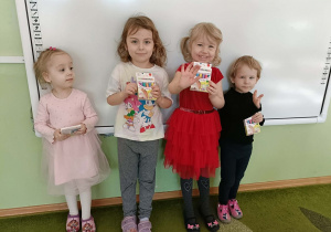 Cztery dziewczynki stoją na tle białej tablicy a w rękach trzymają prezenty.