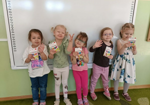 Pięć dziewczynek stoi na tle białej tablicy z prezentami w dłoniach.
