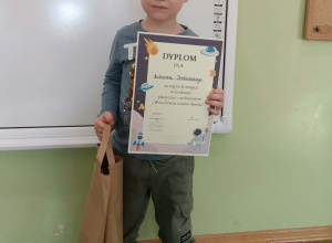 Chłopiec stoi z dyplomem i nagrodą na tle białej tablicy.