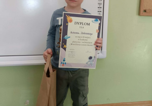 Chłopiec stoi z dyplomem i nagrodą na tle białej tablicy.