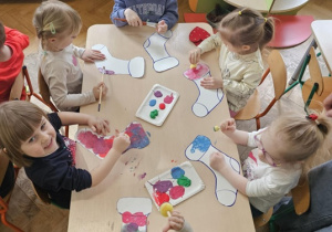 Szóstka dzieci siedzi wokół stolika i maluje farbami szablon skarpetki.