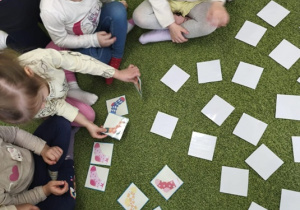 Na zielonym dywanie leżą karty do gry memory z kolorowymi skarpetkami. Wokół siedzą dzieci.