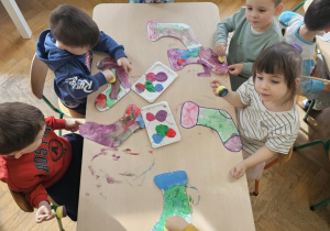 Szóstka dzieci siedzi wokół stolika i maluje farbami szablon skarpetki.