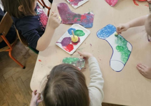 Dzieci siedzą wokół stolika i malują farbami szablon skarpetki.