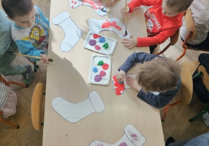 Piątka dzieci siedzi wokół stolika i maluje farbami szablon skarpetki.