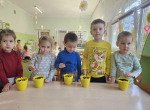 Przy stoliku stoi piątka dzieci. Na stoliku stoją żółte doniczki, w których dzieci sieją owies.