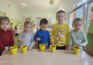 Przy stoliku stoi piątka dzieci. Na stoliku stoją żółte doniczki, w których dzieci sieją owies.