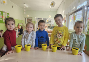 Przy stoliku stoi piątka dzieci. Na stoliku stoją żółte doniczki, w których dzieci sieją owies.