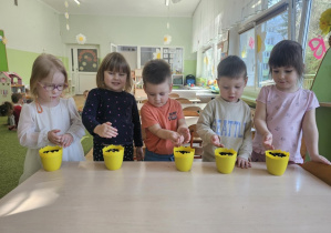 Przy stoliku stoi piątka dzieci. Na stoliku stoją żółte doniczki, w których dzieci sieją owies.
