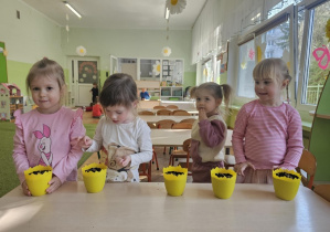 Przy stoliku stoi czwórka dzieci. Na stoliku stoją żółte doniczki, w których dzieci sieją owies.
