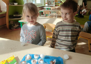 Dwójka dzieci siedzi przy stoliku. Na stoliku leżą pojemniczki z watą i rzeżucha w torebeczkach.