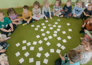 Na zielonym dywanie leżą białe karty. Dzieci siedzą wokół kart.